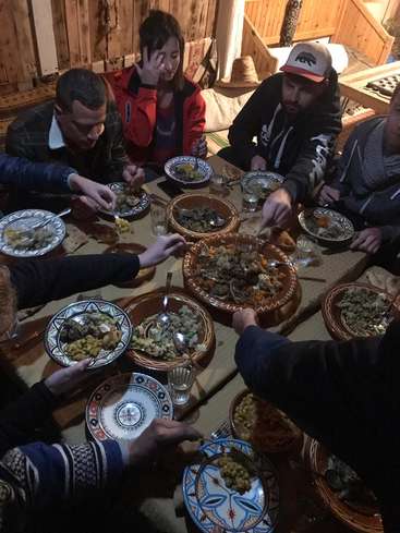 Das Bild zeigt eine Gruppe von Menschen, die um einen Tisch versammelt sind und gemeinsam eine Mahlzeit genießen, wobei der Schwerpunkt auf dem Essen und der warmen Atmosphäre liegt.