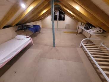 This attic room features a slanted wooden ceiling, two single bed frames, tables with supplies, simple lighting, and minimalistic furnishings, creating a basic, functional space.