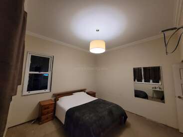This image shows a simple bedroom with beige walls, a double bed, two wooden nightstands, a ceiling light, a window, and a large mirror.