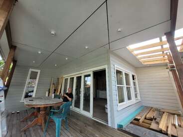 A man sits at a round wooden table on a partially covered porch of a house under construction, surrounded by tools, wood, and unfinished materials.