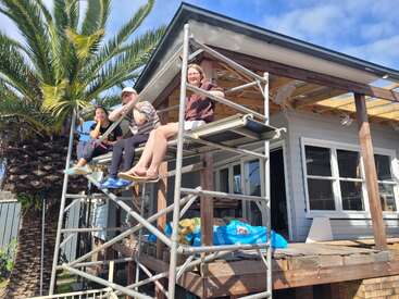 Three people are sitting on scaffolding, smiling and relaxed, beside a house under renovation. A palm tree is nearby, and the sky is partly cloudy.
