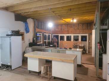 The image shows a kitchen under renovation. Exposed ceiling beams, unfinished walls, construction materials, and tools are visible. A person stands near the windows, working.