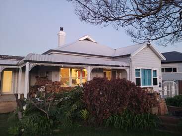 A charming white house with a wraparound porch, decorative trim, blue curtains, and a sloped roof, surrounded by lush greenery and a large overhanging tree.