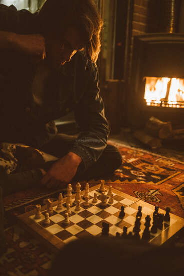 Ein Mann sitzt in einem schwach beleuchteten Raum mit einem Teppich und einem Holzofen auf dem Boden neben einem Kamin und studiert aufmerksam ein Schachbrett mit Figuren, die für ein Spiel aufgestellt sind.