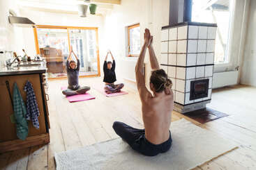 Das Bild zeigt drei Personen, die in einem Raum mit weißen Wänden und Holzboden auf Yogamatten sitzen und ihre Arme in einer Yogapose erhoben haben.