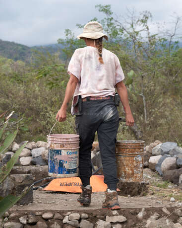 Eine Person geht draußen spazieren, trägt zwei schwere Eimer, ist in robuste Kleidung und Stiefel gekleidet, trägt einen Sonnenhut und ist von Felsen und grüner Vegetation umgeben.