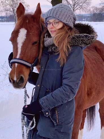 Eine Frau mit Brille, grauer Wintermütze und blauem Mantel steht draußen im Schnee und umarmt liebevoll ein braunes Pferd mit einem weißen Streifen.
