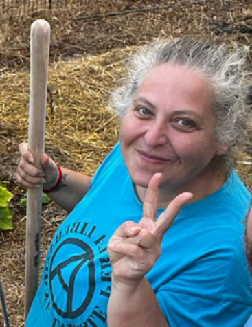 Una mujer de pelo canoso sonríe afectuosamente mientras sostiene un palo de madera y hace el signo de la paz al aire libre, vestida con una camiseta turquesa con un logotipo circular.