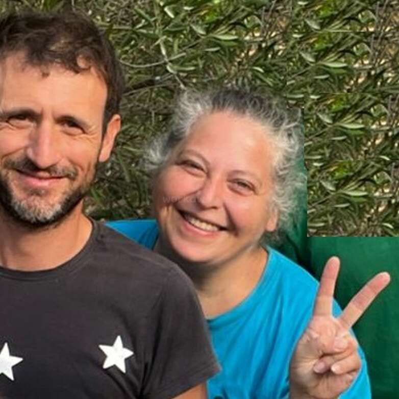 Dos personas están de pie al aire libre, sonriendo cariñosamente a la cámara. La mujer sostiene un signo de la paz, y detrás de ellas hay un fondo de vegetación natural y frondosa.