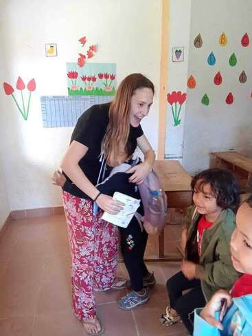 Una profesora sonríe y abraza a un niño en una colorida aula decorada con flores y abecedarios, mientras otros niños felices interactúan cerca. Ambiente cálido.