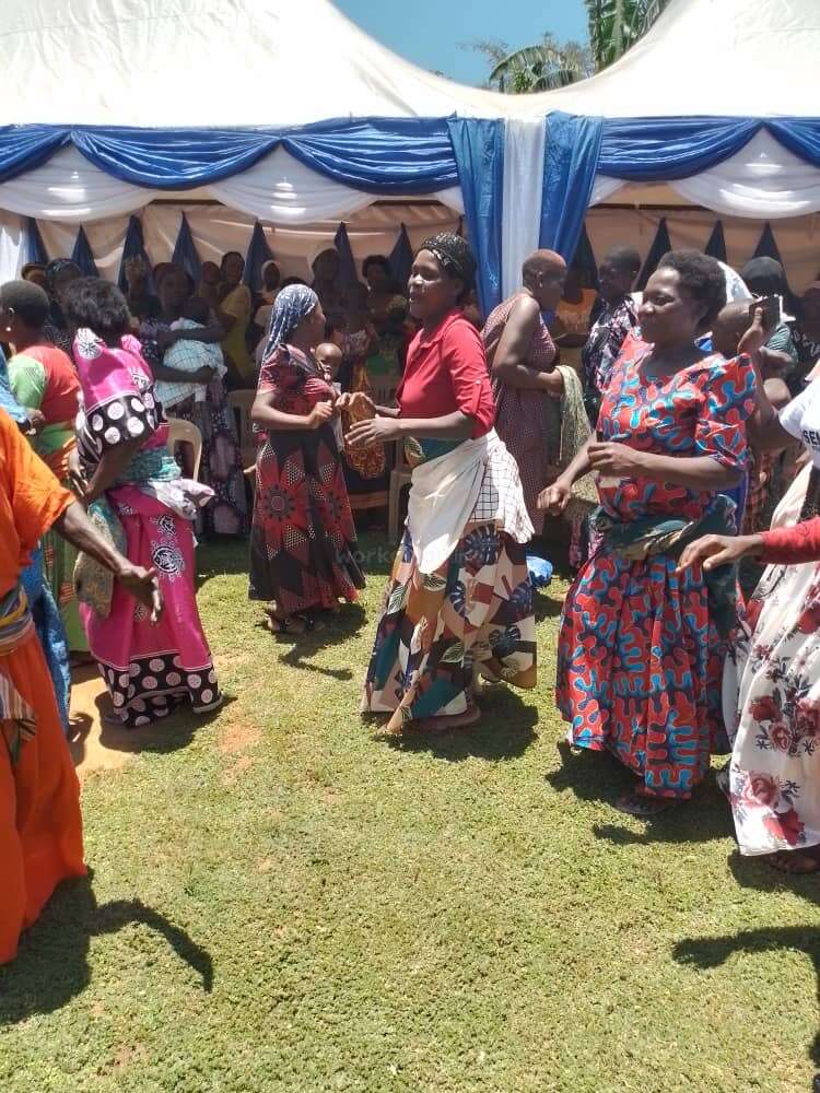 A imagem retrata uma animada reunião de mulheres ao ar livre, vestidas com roupas vibrantes, dançando em um gramado exuberante sob uma tenda branca adornada com cortinas azuis e brancas.