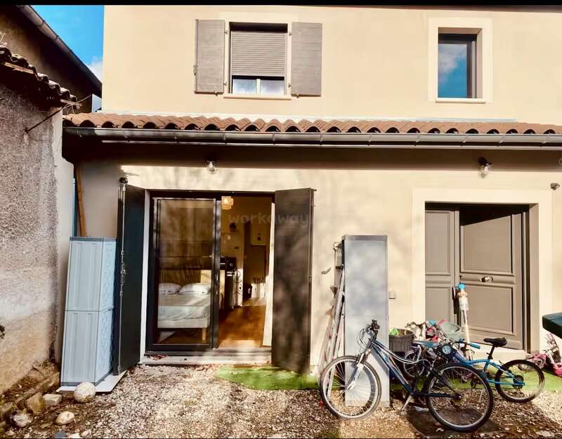 Cette image montre une petite maison à deux étages aux murs couleur crème, avec des volets et des portes gris. Des vélos, des armoires de rangement et divers objets sont disposés dans la cour.