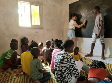 A group of children sits on a mat in a classroom, attentively watching a woman and a boy writing on a blackboard while learning together.