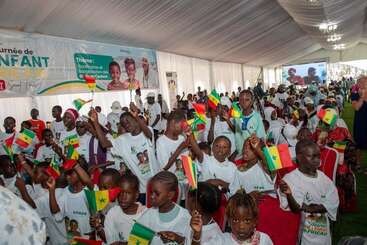 A large group of children are celebrating in a decorated tent, waving Senegalese flags, wearing white shirts, and participating in what appears to be a cultural or national event.