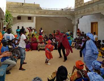 A lively group gathers in a courtyard, with colorful traditional clothing. People joyfully dance, clap, and watch, celebrating together in a friendly, festive atmosphere.