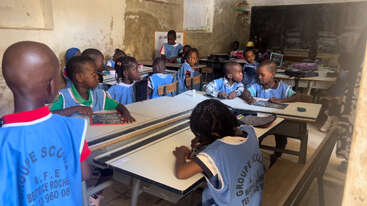 A classroom filled with young children wearing blue uniforms, sitting at desks, engaging in activities. The room has worn walls, a chalkboard, and attentive students.