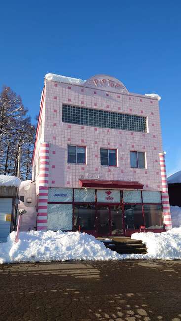 Ein dreistöckiges rosafarbenes Gebäude mit dem Namen "PIPTOP", mit gestreiften Säulen und aufgetürmtem Schnee. Strahlend blauer Himmel, umgeben von Bäumen, japanische Wörter an den Fenstern.