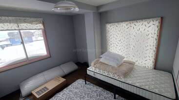 Ein einfaches Schlafzimmer mit zwei Einzelbetten, einer Matratze auf dem Boden, gestapeltem Bettzeug, gemusterten Vorhängen, einem Fenster, durch das man draußen Schnee sieht, und einer Kiste auf dem Boden.