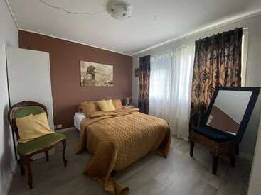 Une chambre douillette avec un lit double, un mur d'accent marron, une parure de lit dorée, des rideaux élégants, une chaise vintage, une table d'appoint, des œuvres d'art et un miroir incliné.
