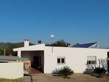 Ein weißes einstöckiges Haus mit Sonnenkollektoren auf dem Dach, eine Terrasse mit Stühlen, zwei kleine Palmen und ein strahlend blauer Himmel im Hintergrund.