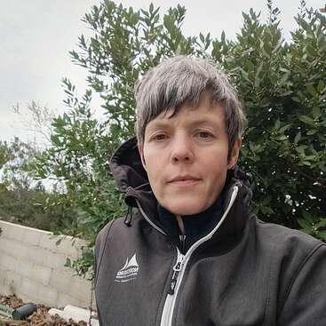 Una persona con el pelo corto y canoso está de pie al aire libre, vestida con una chaqueta oscura de cuello alto. Al fondo se ve vegetación verde y un muro bajo de piedra.