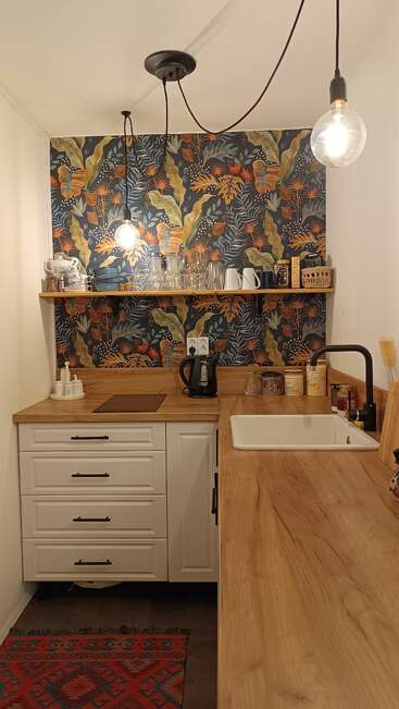 This cozy kitchen features wooden countertops, white cabinets, a vibrant botanical wallpaper, open shelving with dishes, hanging light bulbs, a kettle, and various kitchen essentials.