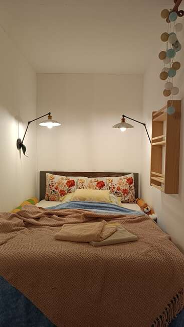 A cozy bed with floral pillows, warm blankets, hanging wall lamps, and decorative lights; towels are neatly folded, creating a welcoming, comfortable, and inviting atmosphere.