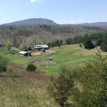 The image depicts a serene rural landscape featuring a house and outbuildings on a hillside, surrounded by trees and mountains in the background under a clear blue sky.