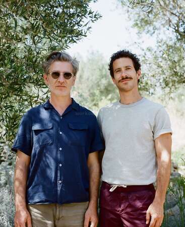 Dois homens estão lado a lado ao ar livre, rodeados por vegetação. Um deles usa óculos escuros e uma camisa azul-marinho; o outro tem bigode, camiseta cinza e calças marrons.