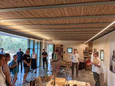 Um grupo de pessoas está reunido em uma sala moderna e bem iluminada, com teto de madeira, ouvindo uma mulher falar. A natureza, estantes de livros e objetos decorativos preenchem o espaço.