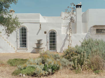 Uma casa branca em estilo mediterrâneo ergue-se sob um céu azul límpido, com janelas em arco, uma chaminé, vasos de plantas, arbustos silvestres e um jardim seco e ensolarado.
