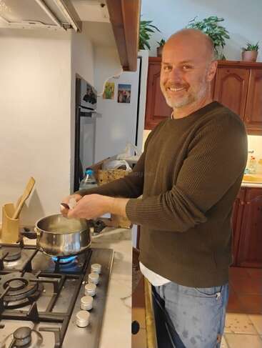 A smiling man stands in a cozy kitchen, cracking an egg into a pot on a gas stove. Plants and wooden cabinets decorate the background.