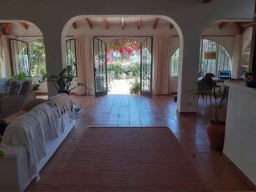 Spacious living room with terracotta floors, arched doorways, and large open windows. Bright natural light, cozy furniture, potted plants, and a charming garden view.