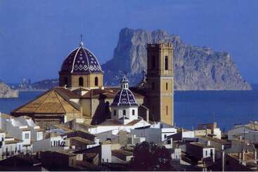 This picturesque coastal town features white buildings, a striking blue-domed church, and a rocky mountain in the background rising majestically above the deep blue sea.