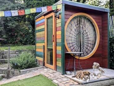 Um pequeno galpão colorido com madeira pintada com listras de arco-íris, uma janela redonda em forma de mandala, bandeiras de oração e dois cachorros relaxados descansando na varanda, rodeados por uma vegetação exuberante.
