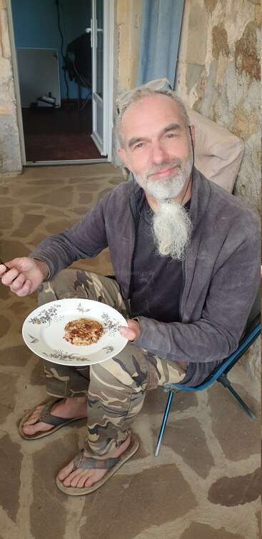 A smiling man with a grey beard sits on a chair outdoors, eating food from a plate. He wears camouflage pants, flip-flops, and glasses on his head.