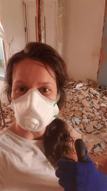A woman wearing a protective mask and blue gloves gives a thumbs-up while standing in a room under renovation, with rubble and broken tiles on the floor.
