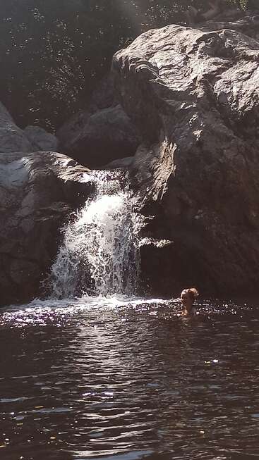 Ein kleiner Wasserfall stürzt über schroffe Felsen in ein dunkles Becken. Das Sonnenlicht glitzert auf dem Wasser, während eine Person in der Nähe der Basis schwimmt. Gelassene Natur.