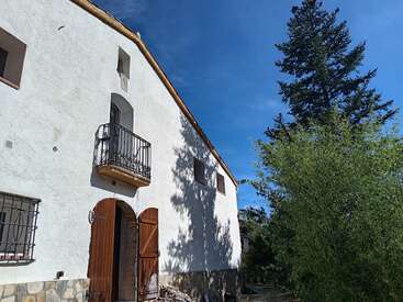 Esta imagen muestra una casa rústica blanca con un pequeño balcón, puertas de madera y ventanas con rejas. Se ven árboles altos y verdes y un cielo azul claro cerca.