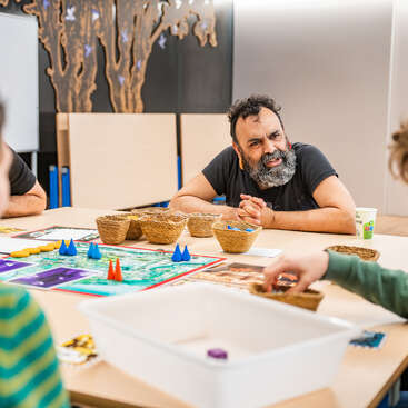 Three people sit around a table, playing a board game together. The atmosphere appears friendly and engaged, with colorful game pieces and baskets visible.