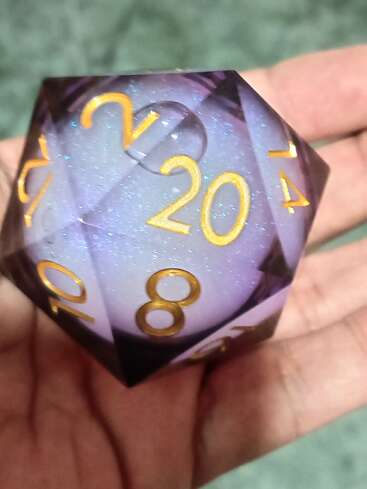 Uma mão segura um grande dado roxo brilhante de vinte faces (d20) com números dourados. O dado tem um núcleo brilhante e translúcido com uma aparência caprichosa e mística.