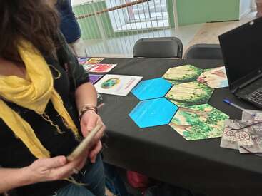 Uma mulher com um lenço amarelo está ao lado de uma mesa com cartões hexagonais com imagens da natureza, folhetos, um laptop e outros materiais coloridos em um evento.