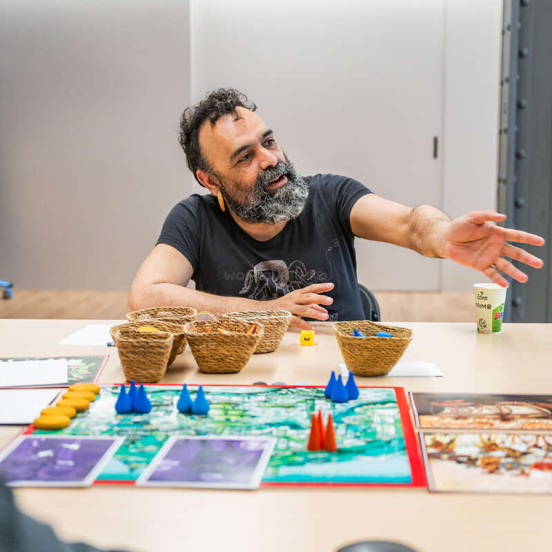Um cara barbudo faz gestos animados enquanto joga um jogo de tabuleiro. Cestas de vime com peças e cartas do jogo estão na mesa. Um copo com bebida está ao lado.