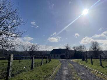 A bright, sunny day illuminates a rural scene with a gravel driveway, green fields, bare trees, fenced pastures, and a quaint house in the distance.