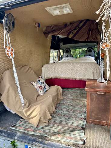 O interior aconchegante desta van conta com um futon revestido de bege, almofadas estampadas, uma cama com roupa de cama geométrica, um tapete tecido, uma mesinha de cabeceira de madeira, cestas de frutas penduradas e um pequeno ventilador.