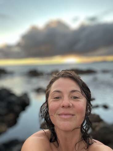 Uma mulher com o cabelo molhado e um piercing no nariz sorri para a câmera, em frente a uma costa rochosa pitoresca com um pôr do sol nublado e espetacular.