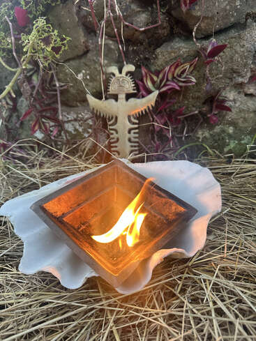 Uma fogueira cerimonial arde dentro de um recipiente de metal, colocado sobre uma grande concha em meio à palha. Atrás dela, um totem tribal se ergue contra uma parede de pedra coberta de plantas.