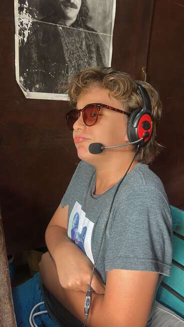 Un jeune homme portant des lunettes de soleil et un casque audio est assis, les bras croisés, l'air détendu. Derrière lui se trouve un portrait noir et blanc usé représentant un homme moustachu.