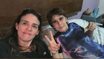 Une femme et un garçon sont assis ensemble sur un lit ; ils sourient à la caméra en faisant le signe de la paix. Le garçon tient un en-cas.
