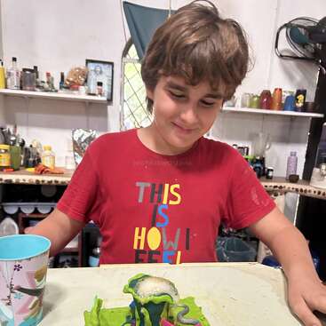 A smiling boy in a red “THIS IS HOW I FEEL” shirt sits at a table, observing a colorful homemade science experiment with bubbling foam and slime.